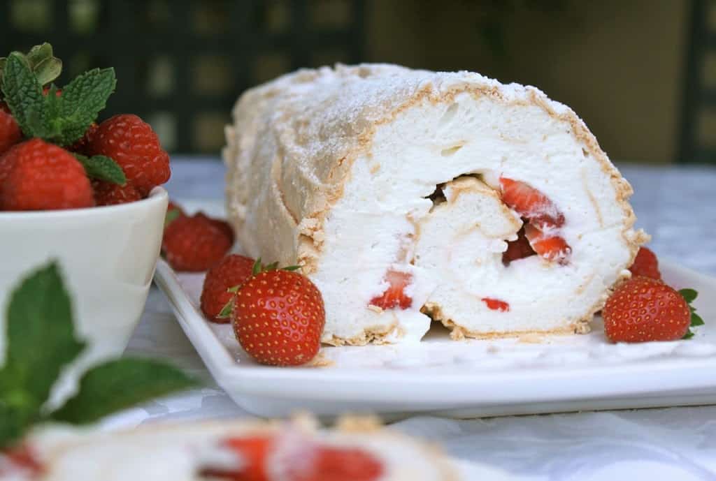 A meringue roulade filled with whipped cream and sliced strawberries, served on a white plate with whole strawberries and mint leaves.