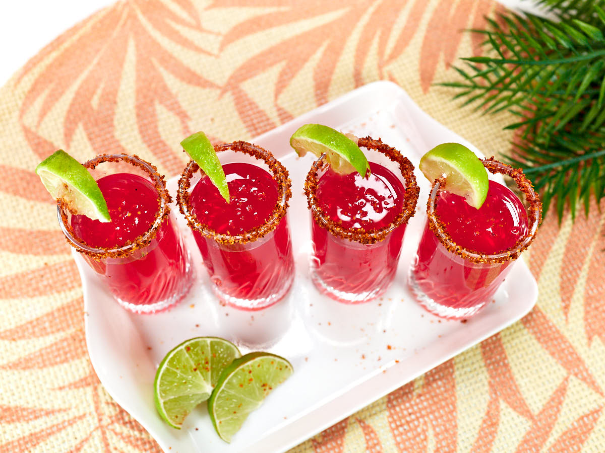 Four red cocktails with chili-rimmed glasses and lime wedges are lined up on a white tray, with extra lime slices beside them on a woven mat.