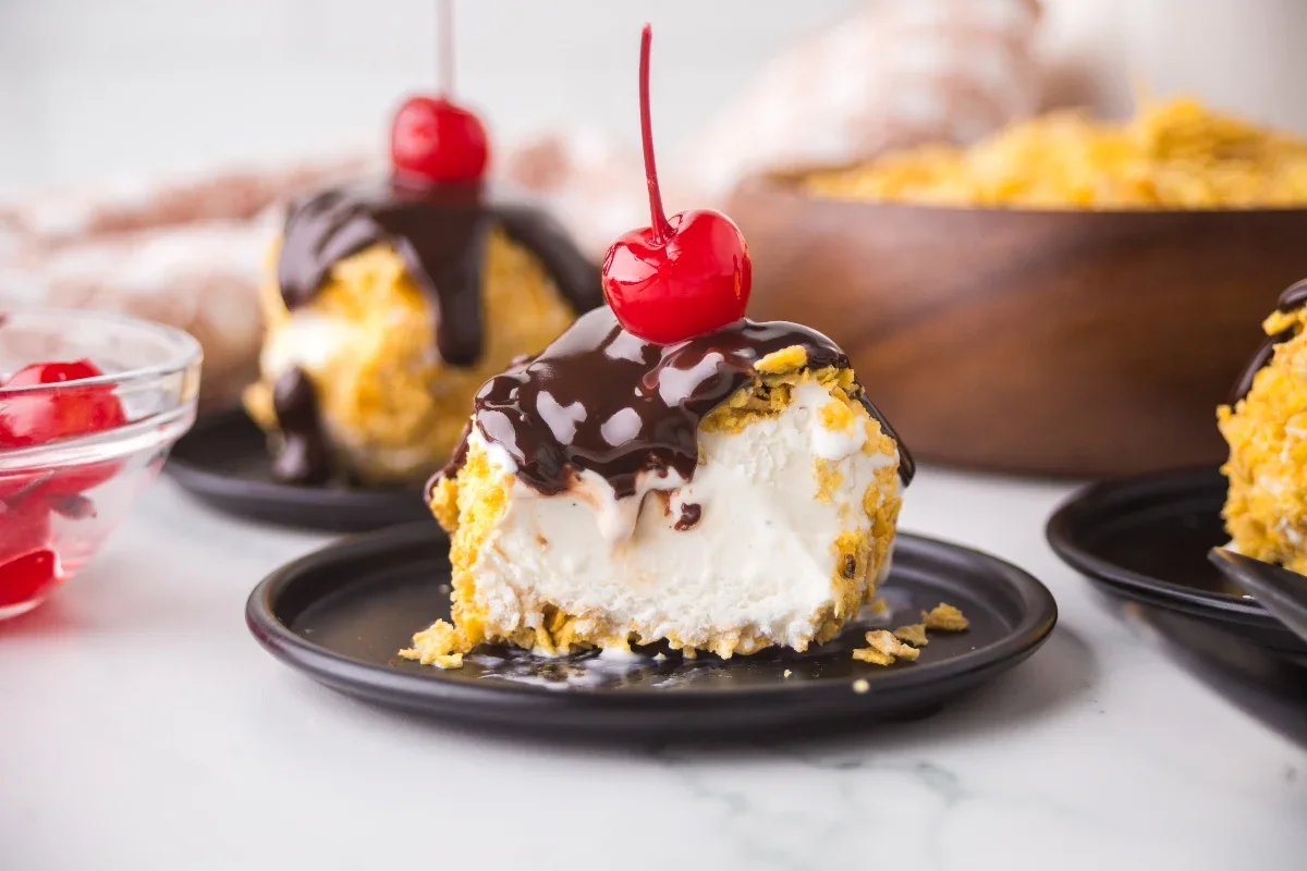A scoop of vanilla ice cream coated in a crunchy shell, topped with chocolate sauce and a cherry, served on a black plate.