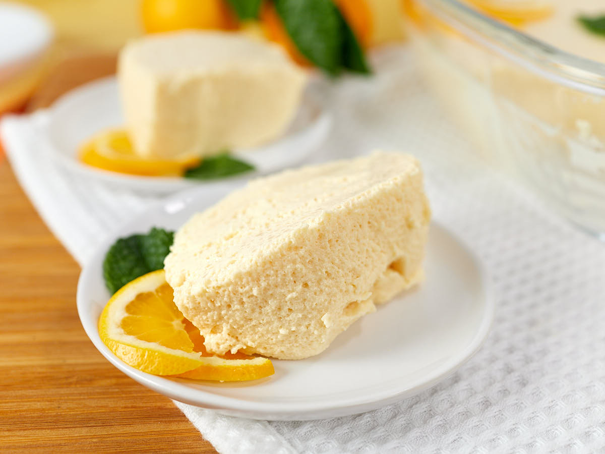 A slice of creamy orange mousse cake on a white plate, garnished with orange slices and mint leaves, with another slice and baking dish in the background.