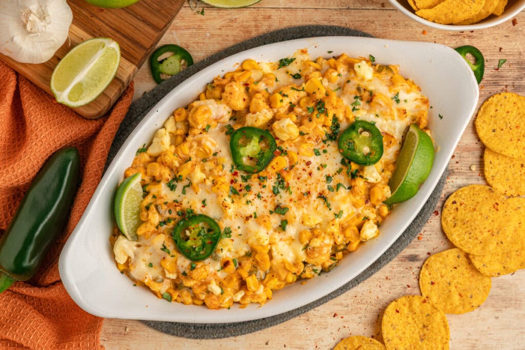 Oval dish filled with cheesy corn dip, garnished with jalapeño slices, lime wedges, and herbs, surrounded by tortilla chips, a lime half, garlic, and a jalapeño pepper.