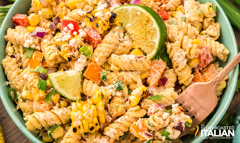 A bowl of pasta salad with rotini, corn, diced vegetables, green onions, feta cheese, and lime wedges, served with a wooden fork.