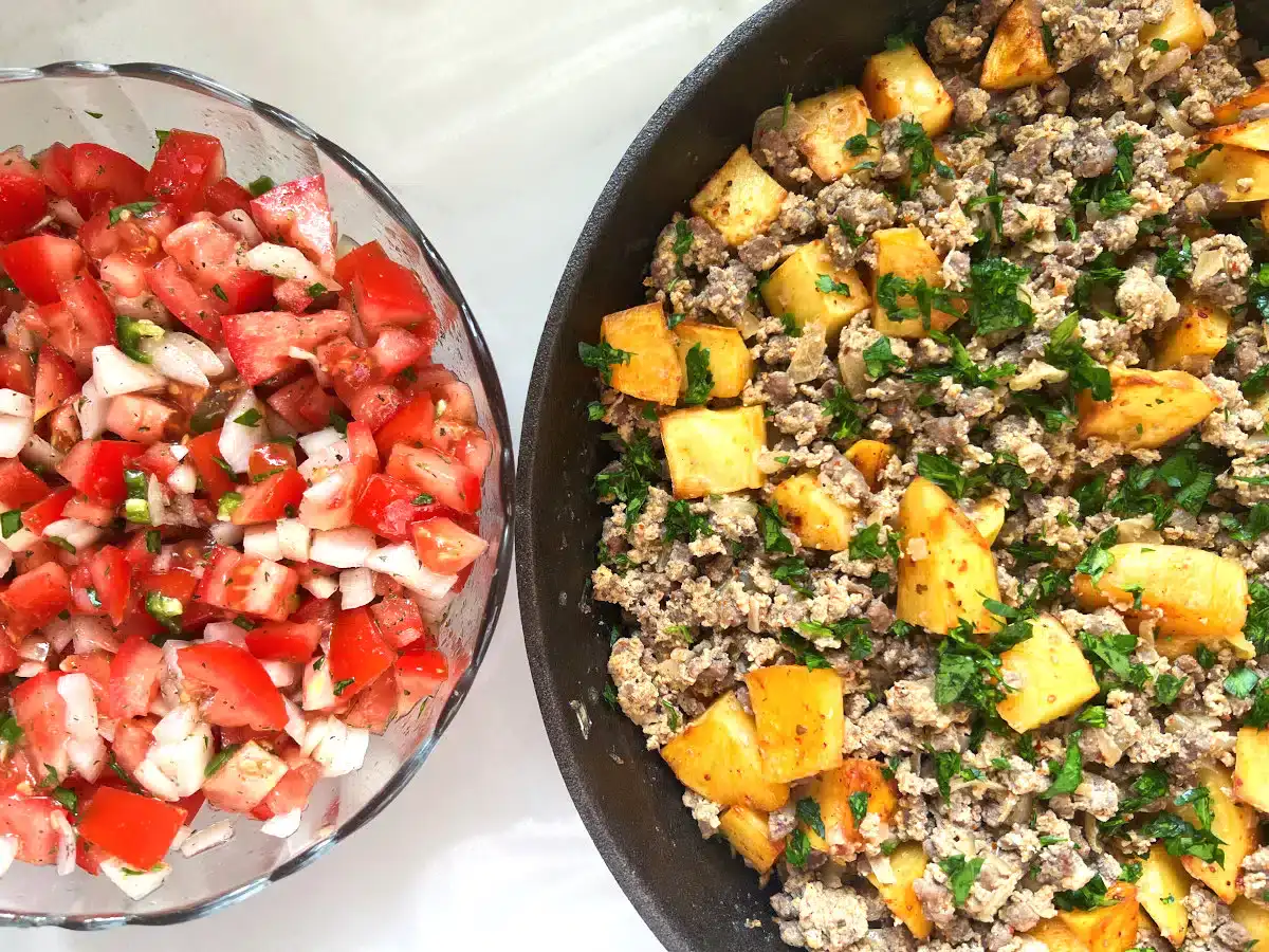 A bowl of tomato and onion salad sits next to a skillet containing ground meat, potatoes, and chopped herbs.