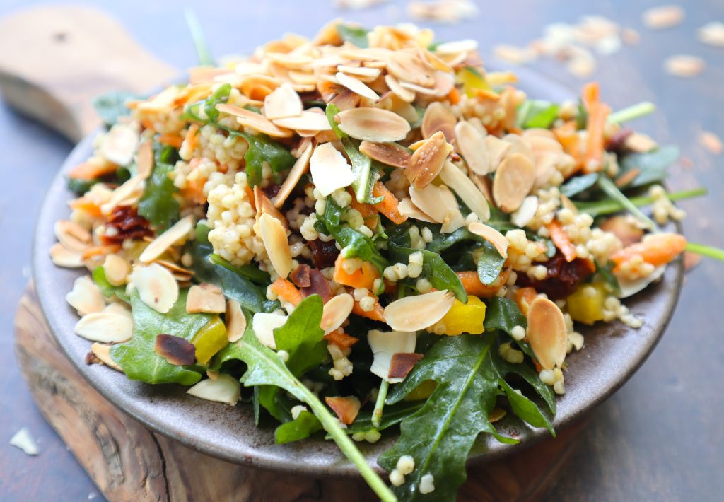 A plate of salad with leafy greens, cooked couscous, shredded vegetables, sun-dried tomatoes, and topped with toasted almond slices.