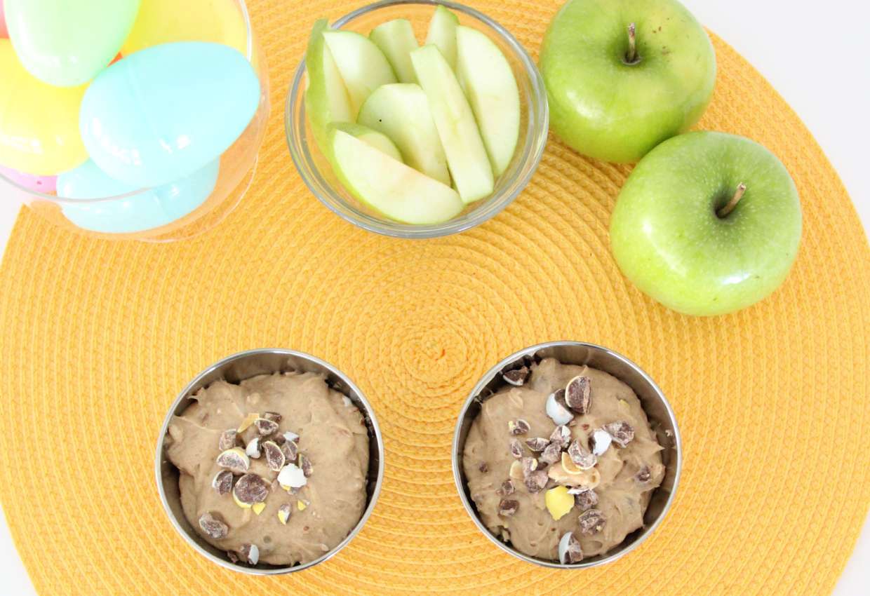Two green apples, a bowl of apple slices, plastic Easter eggs, and two bowls of dessert topped with crushed chocolate candies are arranged on a yellow mat.