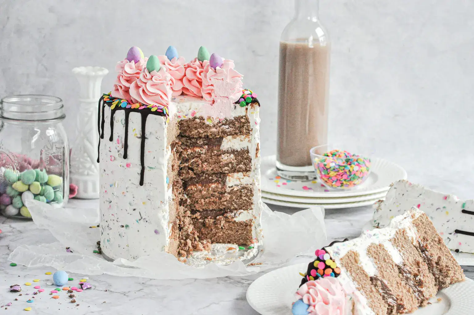 A tall, multi-layered cake with white and pink frosting, chocolate drizzle, and colorful sprinkles, sits partially sliced on a table with plates, a bottle, and jars of candy nearby.