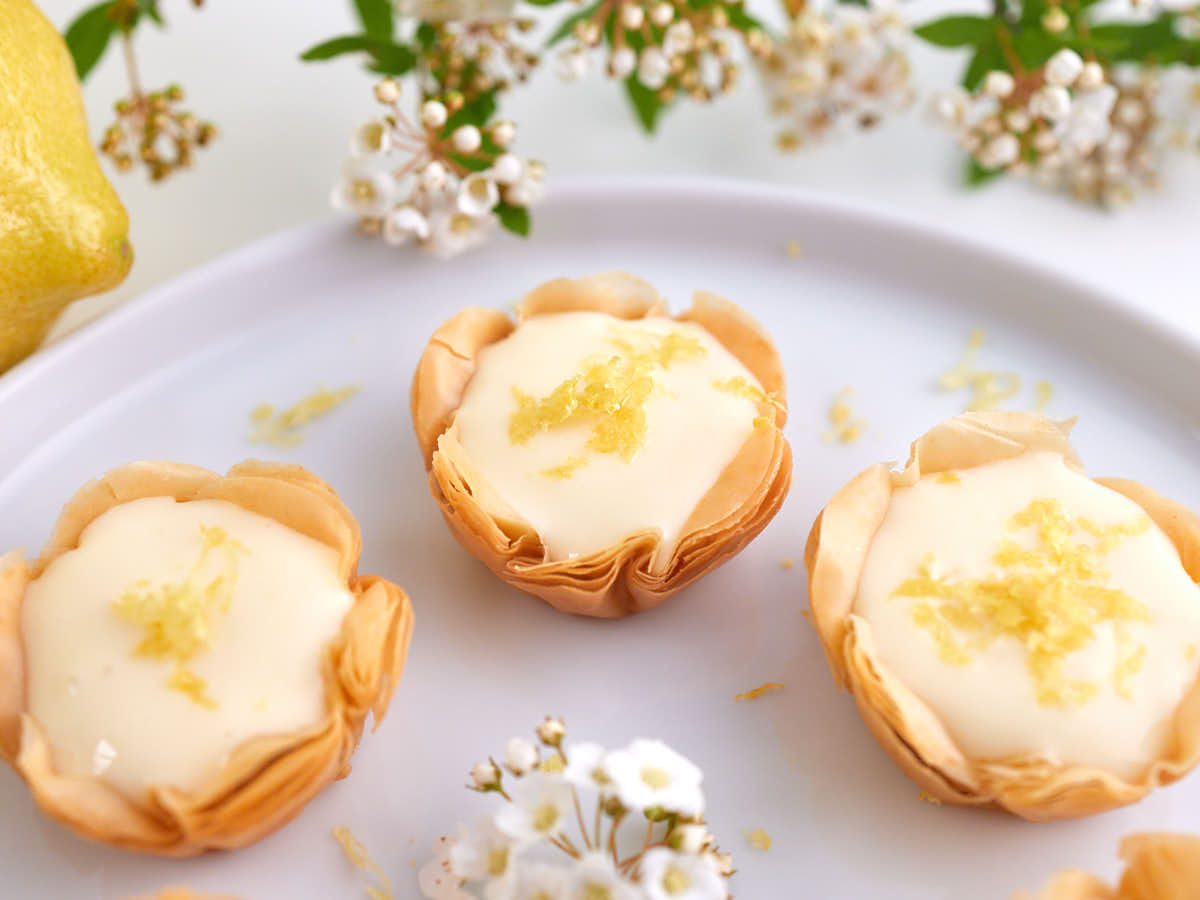 Three mini lemon tarts with flaky pastry shells and creamy filling, topped with lemon zest, are arranged on a white plate with small white flowers nearby.
