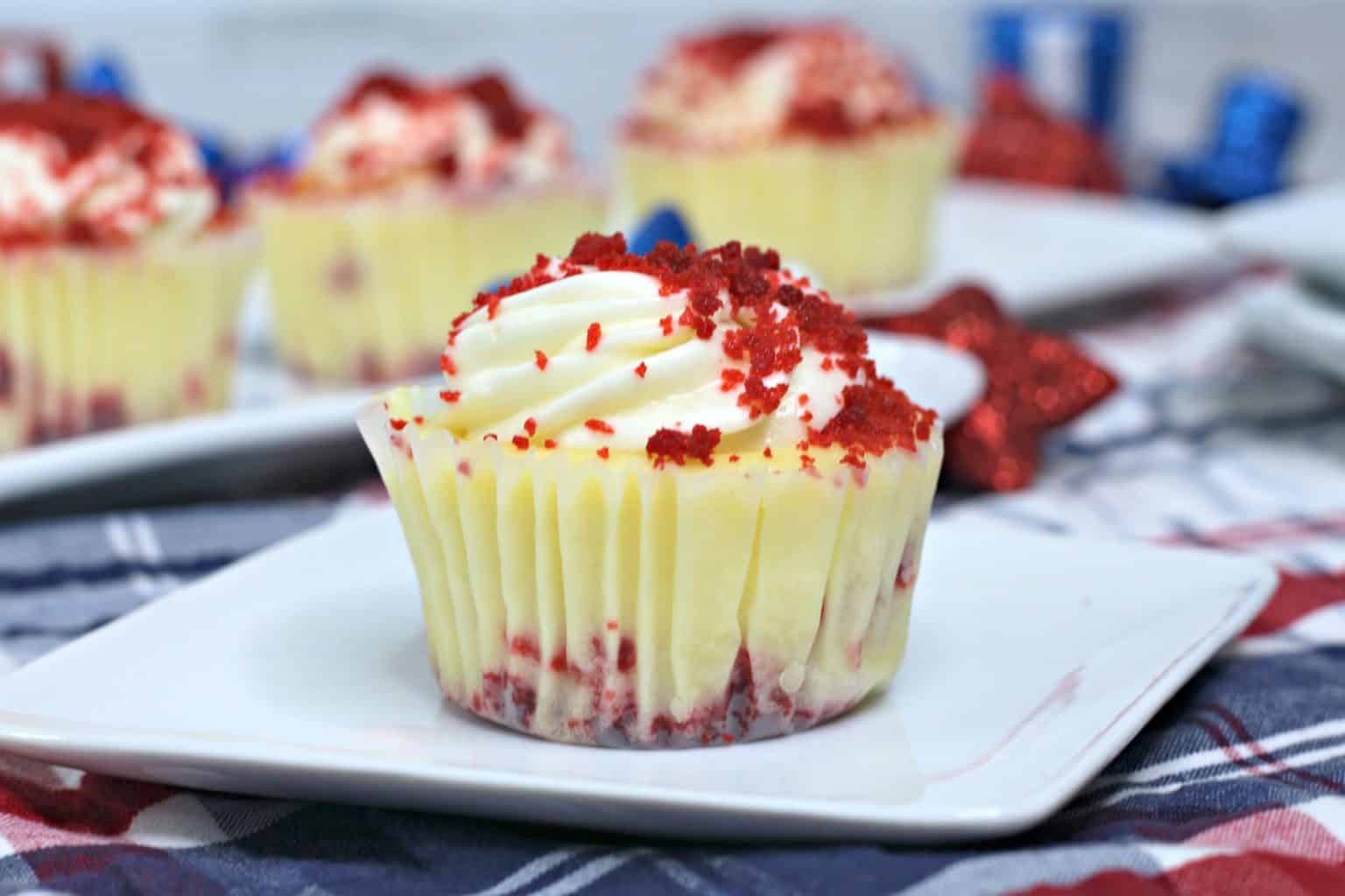 A red velvet cupcake with cream cheese frosting and red crumbs sits on a white plate, with more cupcakes in the background.