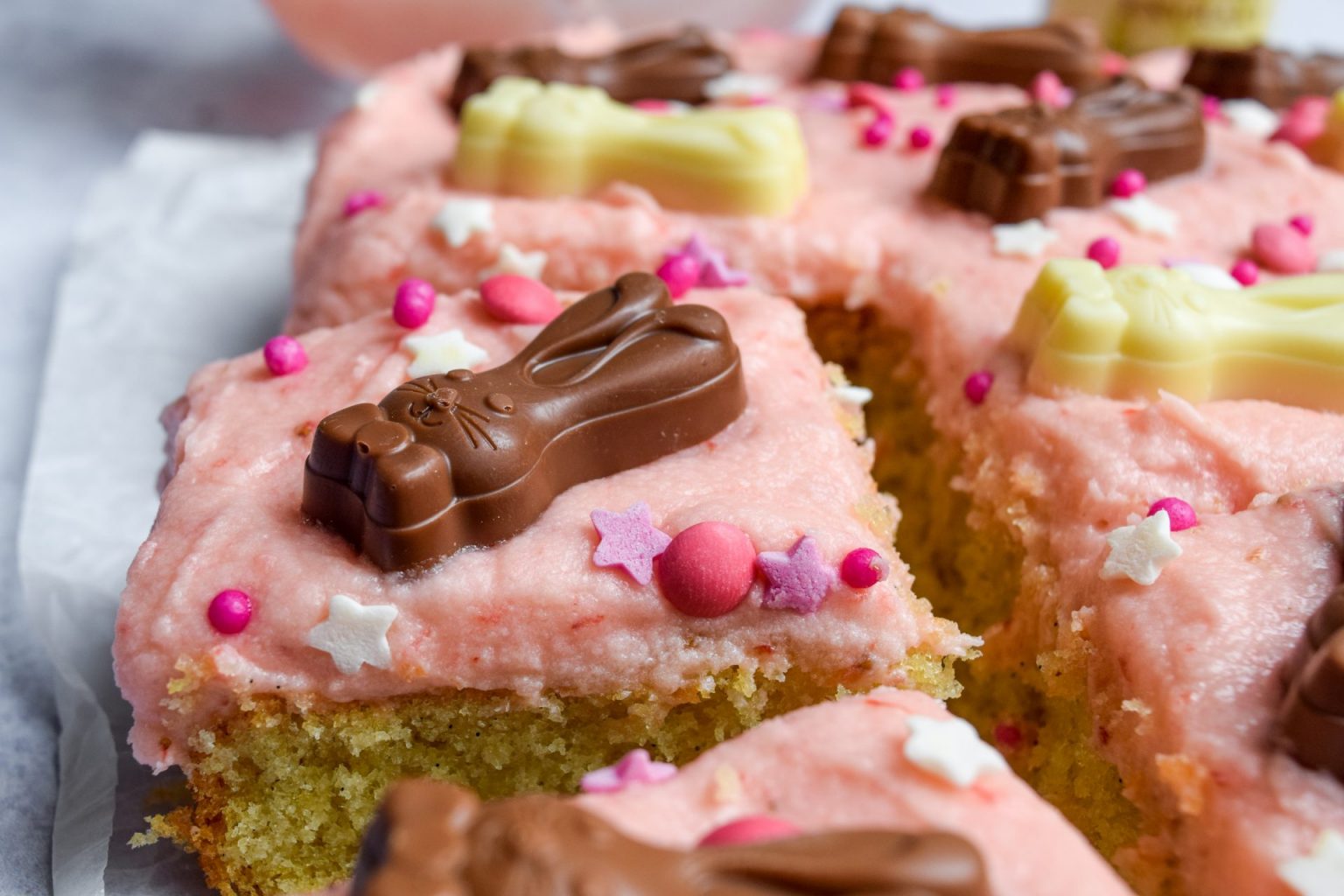 A close-up of a cake with pink frosting, decorated with chocolate and white chocolate bunny shapes, pink and white star sprinkles, and round candy pieces.