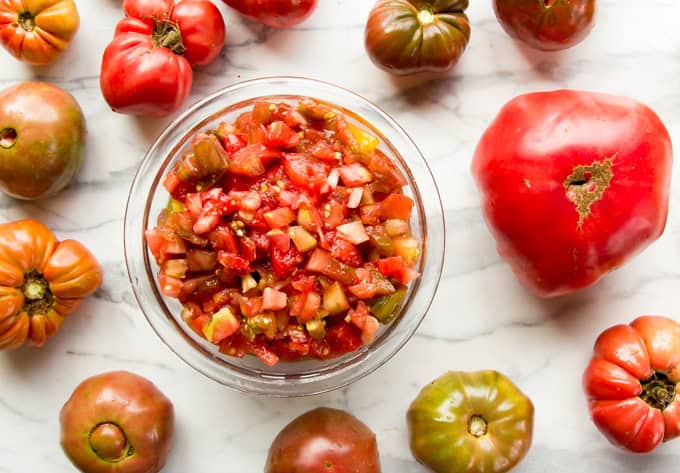 A glass bowl of chunky tomato salsa surrounded by whole heirloom tomatoes on a white marble surface.