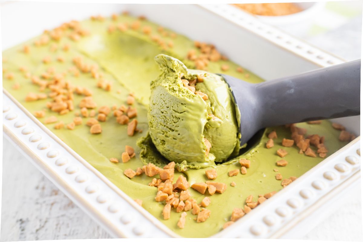 A close-up of green ice cream being scooped from a rectangular dish, topped with small caramel toffee pieces.