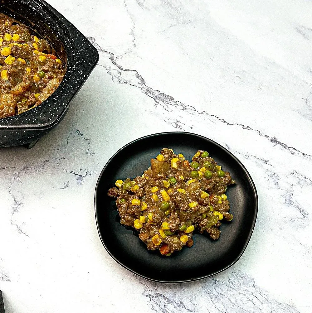 A serving of ground beef casserole with corn, peas, and diced potatoes on a black plate, next to a pot with more of the same casserole on a white marble surface.