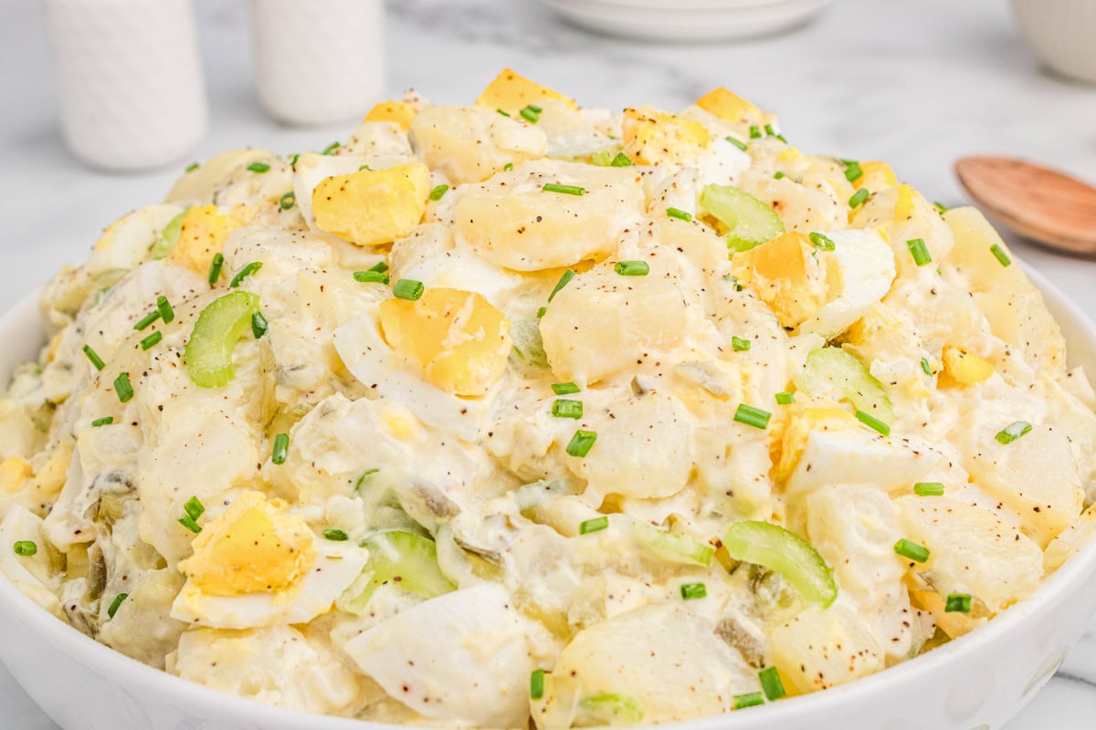A bowl of potato salad with sliced boiled eggs, celery, and chopped chives, mixed with creamy dressing and sprinkled with black pepper.