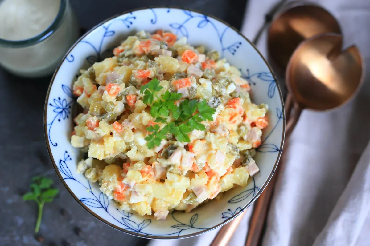 A bowl of potato salad with diced vegetables and mayonnaise, garnished with parsley. Copper serving utensils are placed beside the bowl.