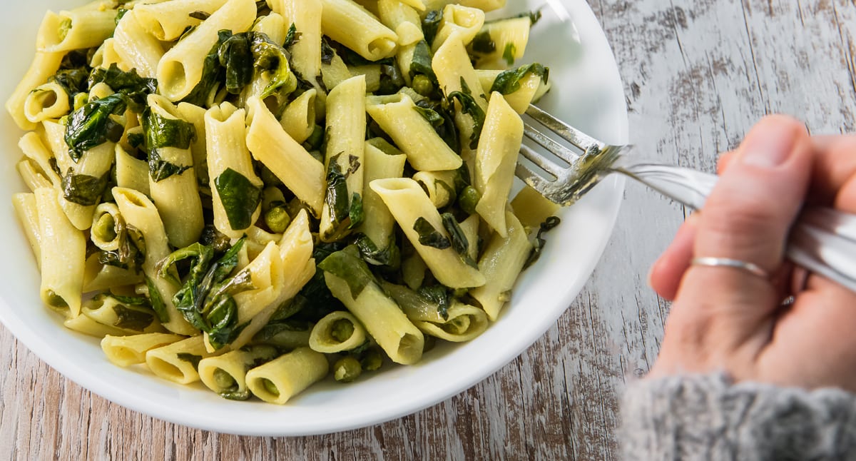 A hand holding a fork is about to eat a bowl of penne pasta mixed with spinach and green peas on a wooden surface.