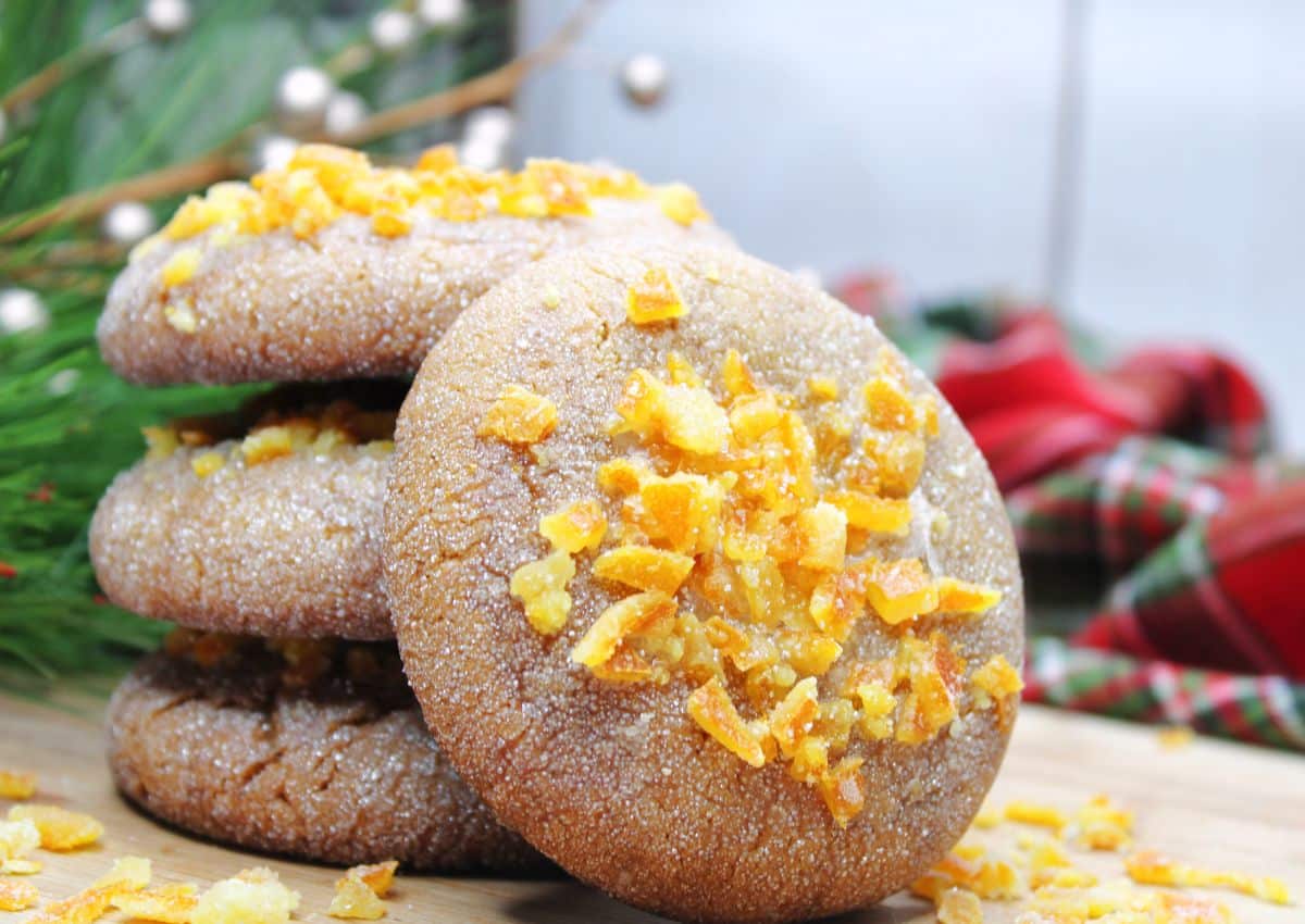 A stack of sugar-coated cookies topped with orange zest pieces on a wooden surface, with holiday decorations in the background.