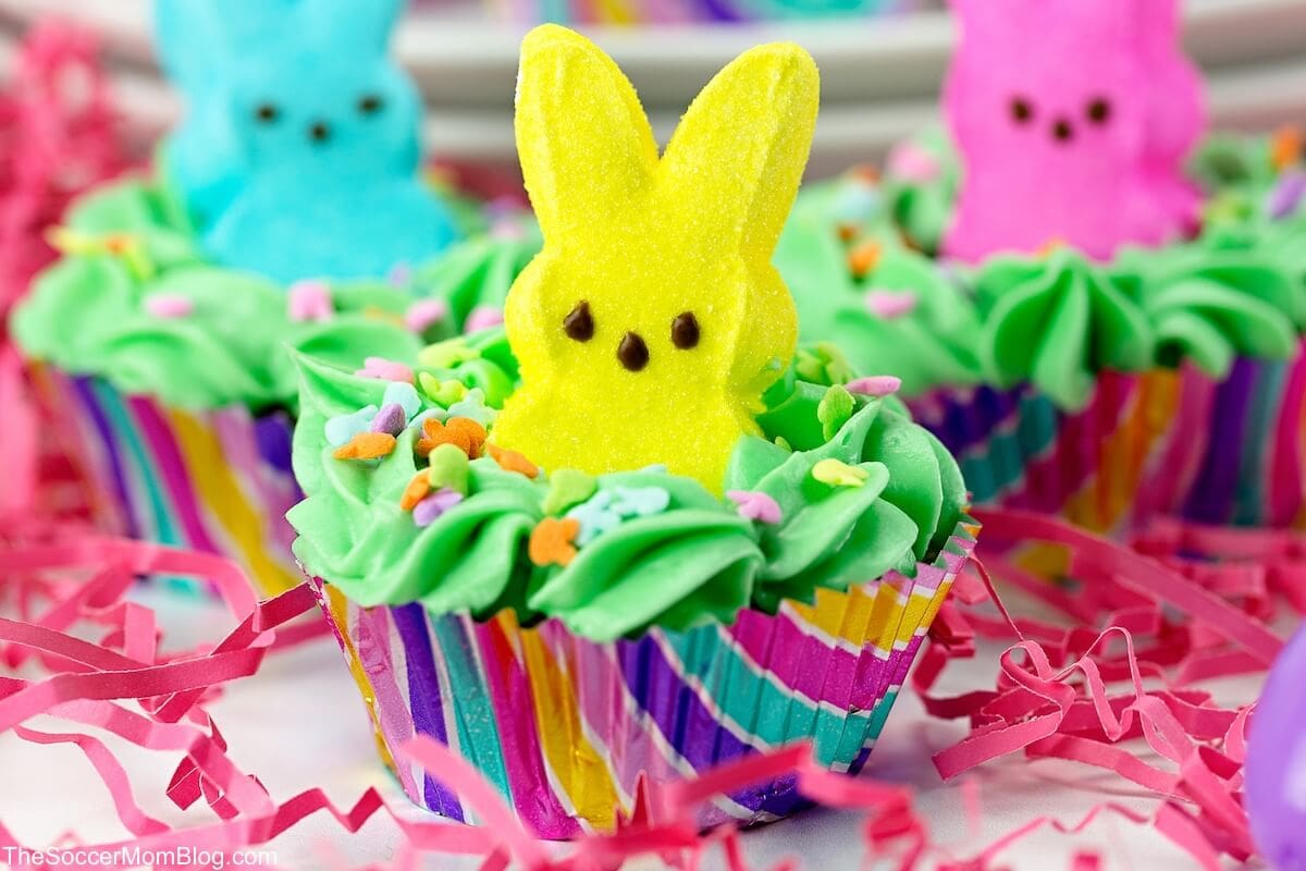 Colorful cupcakes topped with green frosting, pastel sprinkles, and a yellow marshmallow bunny, with more decorated cupcakes in the background and pink paper shreds underneath.