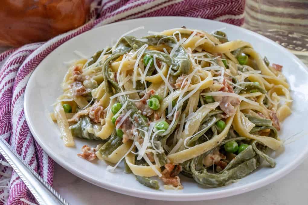 A plate of fettuccine pasta with green peas, bacon pieces, and grated cheese, served on a white dish with a striped cloth napkin in the background.