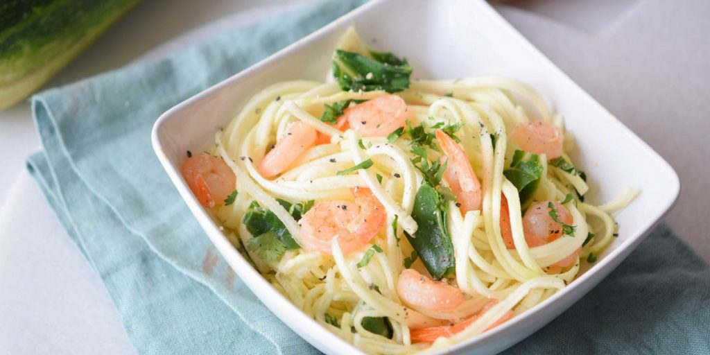 A square white bowl filled with pasta, shrimp, and chopped green vegetables sits on a light blue cloth.