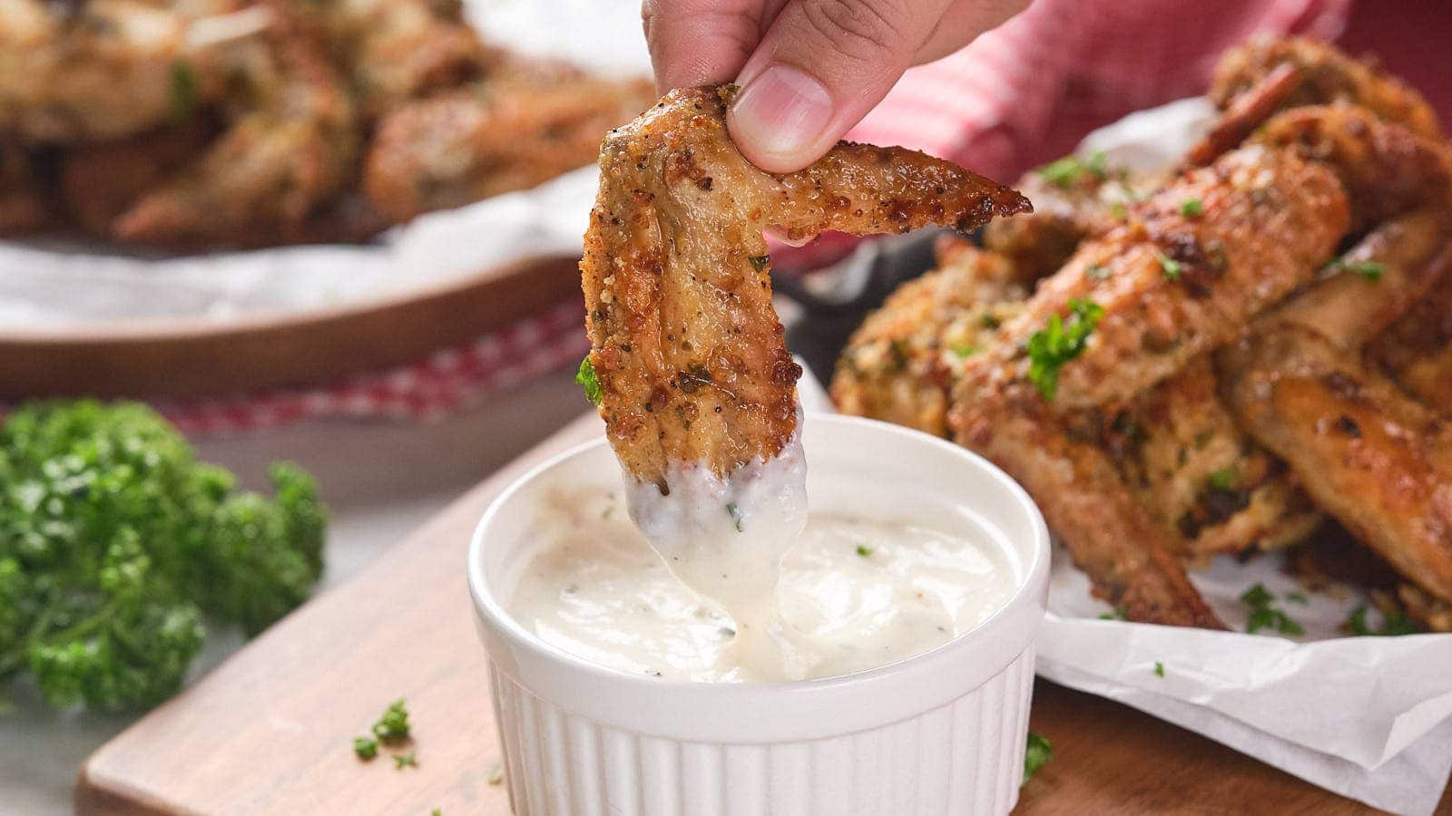A hand dipping a seasoned chicken wing into a small bowl of creamy white sauce, with more wings and herbs visible in the background.