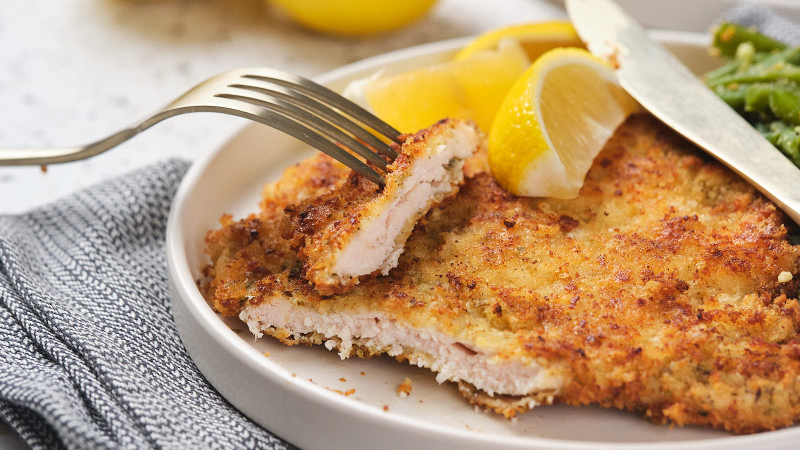 A breaded and cooked chicken cutlet on a plate with lemon wedges and a fork holding a piece, next to green beans.