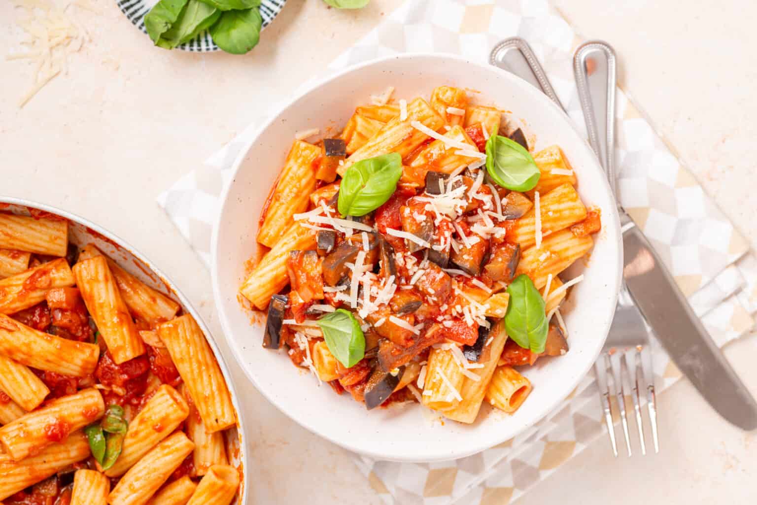 A bowl of rigatoni pasta with tomato sauce, eggplant, grated cheese, and basil leaves, placed next to a pan of pasta and cutlery on a patterned napkin.