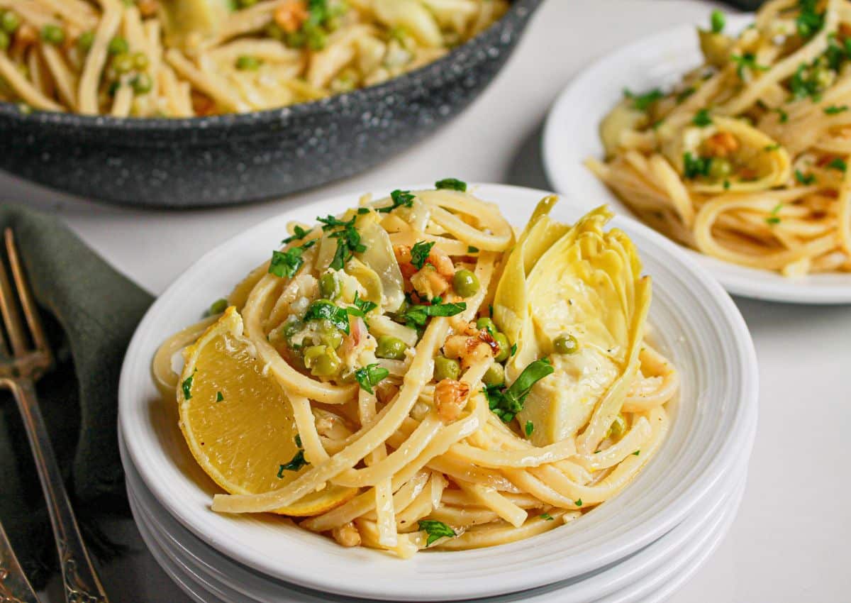 A plate of linguine pasta topped with artichoke hearts, lemon slices, chopped parsley, and nuts, with more pasta and a second plate in the background.