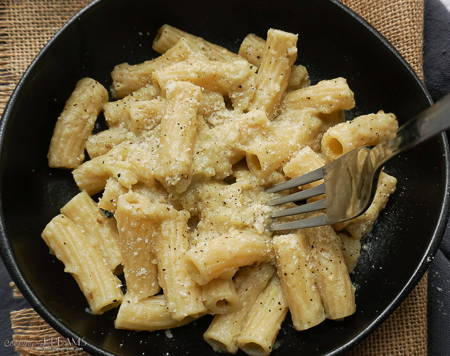 A black bowl filled with rigatoni pasta coated in a creamy sauce, sprinkled with grated cheese and black pepper, with a fork resting on the pasta.
