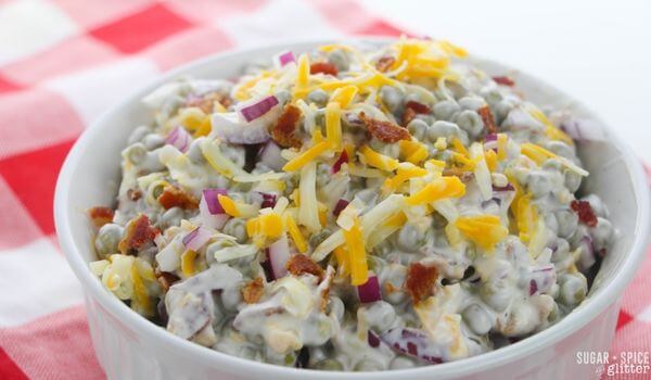 A white bowl filled with creamy pea salad topped with shredded cheddar cheese, chopped red onions, and bacon bits sits on a red and white checkered tablecloth.