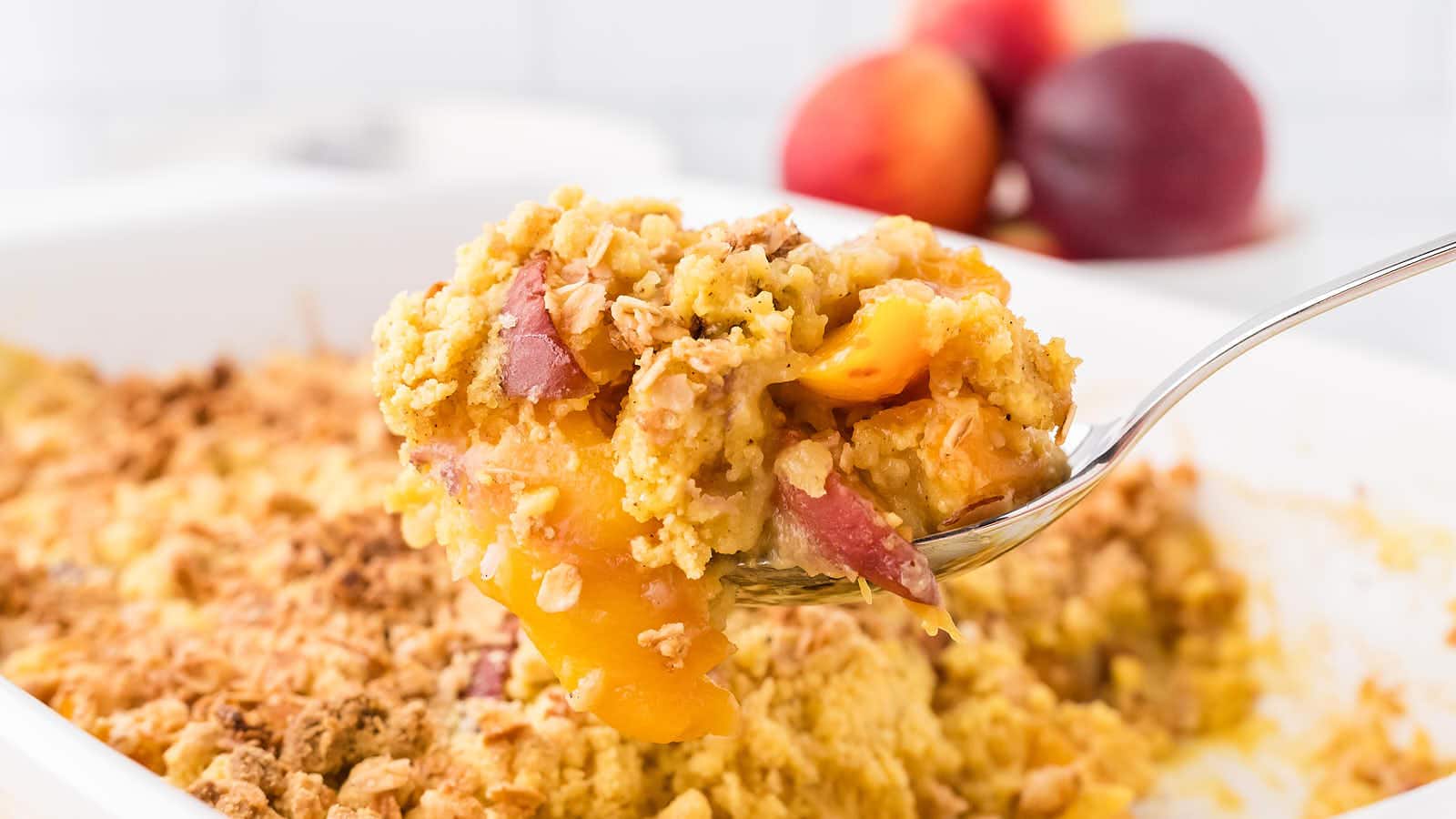 A spoonful of peach crumble is held above a baking dish, with fresh peaches blurred in the background.