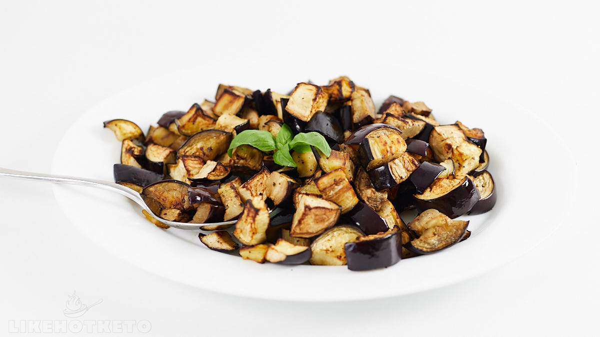 A white plate with roasted, diced eggplant garnished with fresh basil, accompanied by a silver fork, on a plain white background.