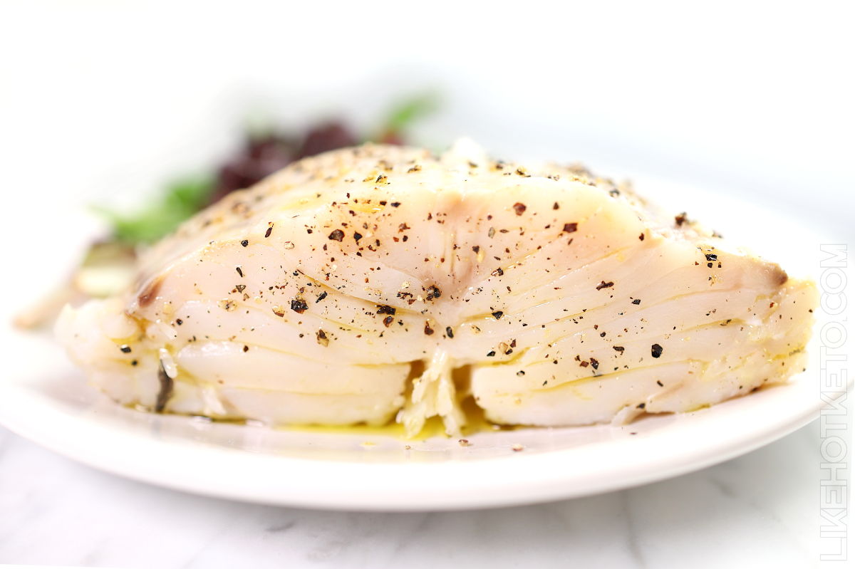 A cooked fillet of white fish seasoned with black pepper and herbs, served on a white plate with a small portion of greens in the background.