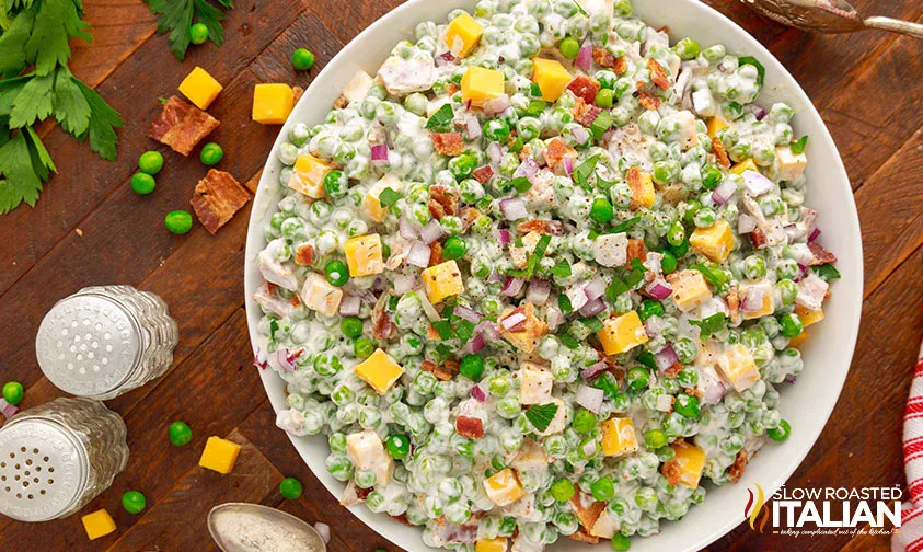 A bowl of pea salad with diced cheddar cheese, bacon pieces, red onion, and a creamy dressing sits on a wooden table with scattered ingredients and salt and pepper shakers nearby.