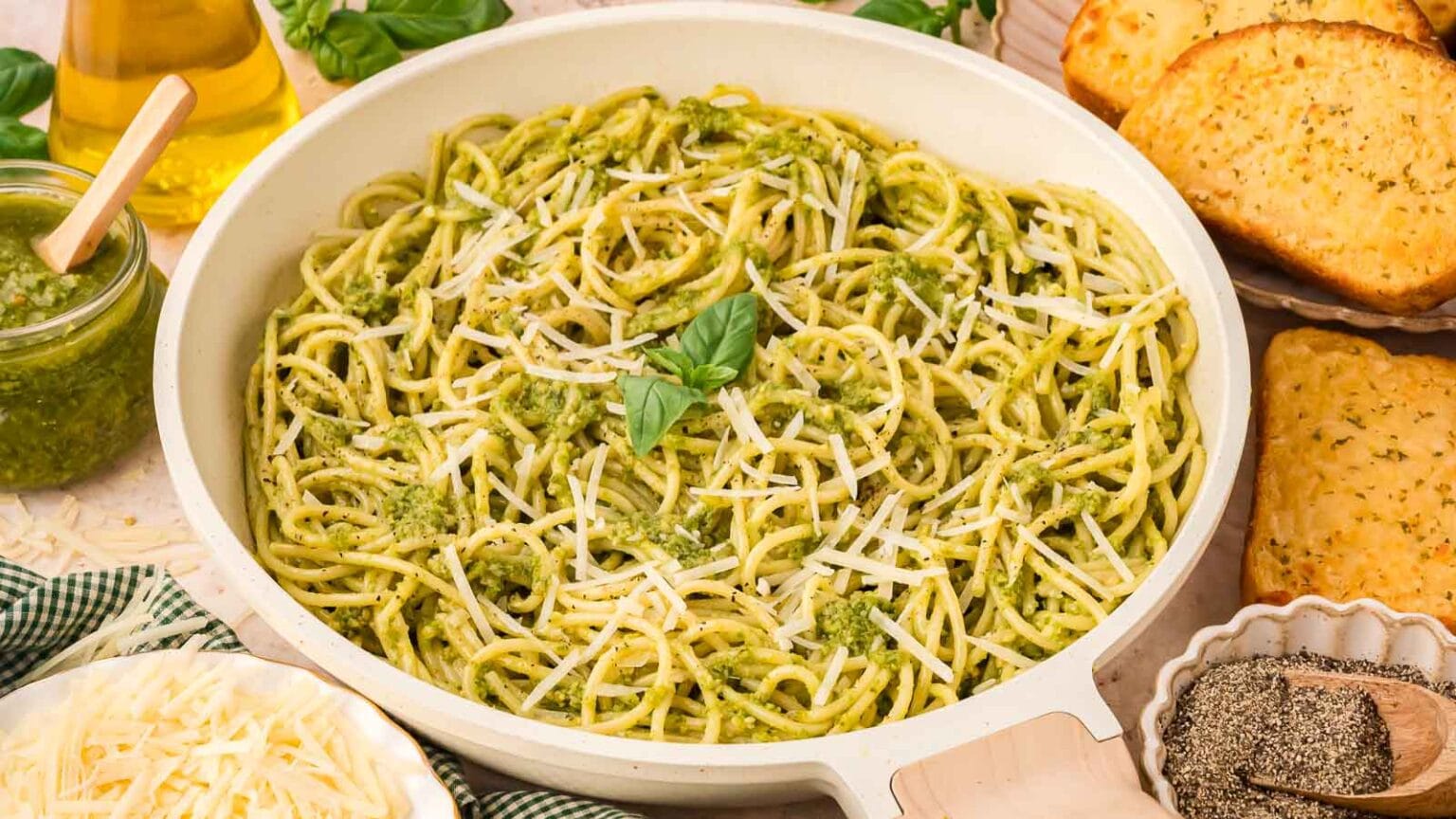 Cooked spaghetti pasta with green pesto sauce and grated cheese in a white pan, surrounded by garlic bread, grated cheese, and a jar of pesto.