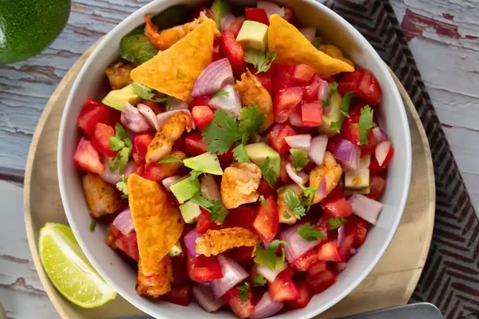 A bowl of salsa with diced tomatoes, onions, avocado, grilled chicken, cilantro, and tortilla chips, served with a lime wedge on the side.