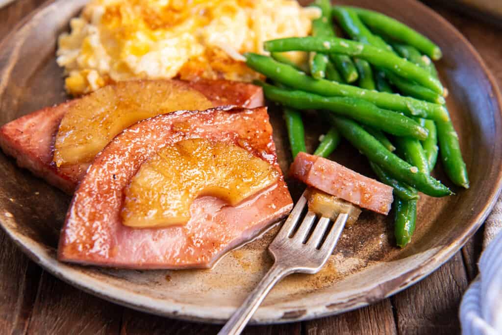 A plate with glazed ham topped with pineapple slices, green beans, and a serving of cheesy potatoes. A fork holds a piece of ham.