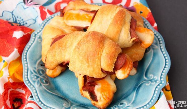 A blue plate holds several baked crescent rolls filled with melted cheese and slices of deli meat, set on a colorful floral napkin.