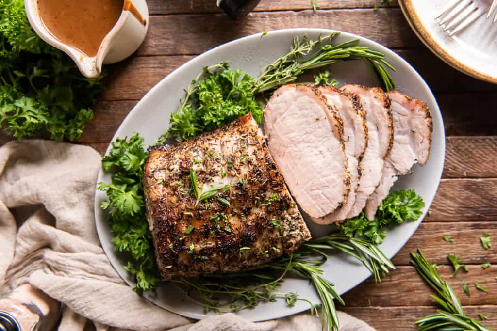 Sliced herb-crusted pork roast on a white platter, garnished with fresh parsley and rosemary, set on a wooden table with a light-colored napkin.