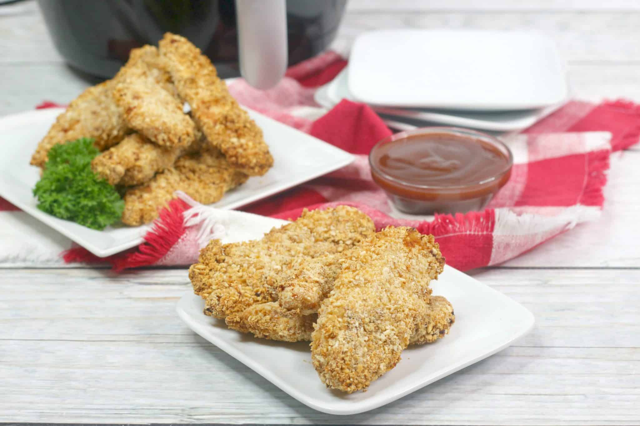 Plates of breaded chicken tenders are served with a side of barbecue sauce, placed on a red and white cloth with an air fryer in the background.