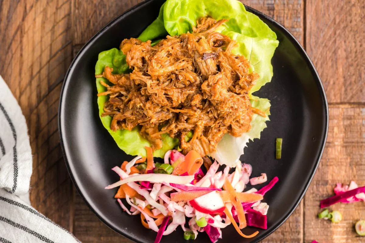 A black plate with shredded meat on lettuce leaves and a side of colorful coleslaw, placed on a wooden table.
