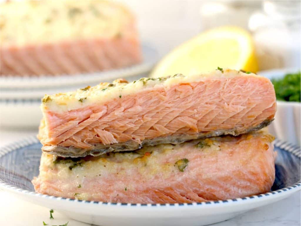 Two pieces of cooked salmon stacked on a white plate with a blue rim, with a lemon wedge and seasoning in the background.