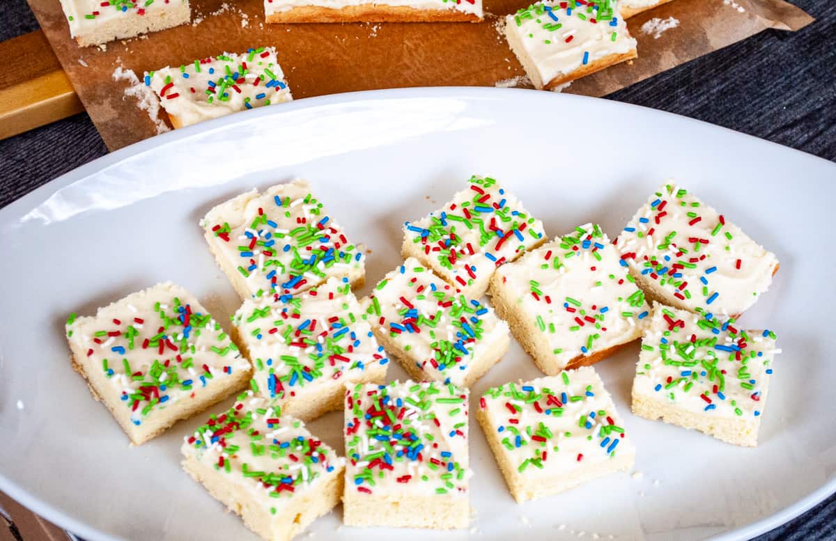 Rectangular sugar cookie bars with white frosting and red, blue, and green sprinkles arranged on a white plate, with more bars visible in the background.