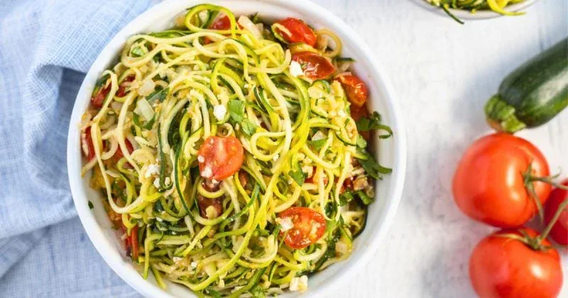 A white bowl filled with zucchini noodles, halved cherry tomatoes, and herbs. Two whole tomatoes and a zucchini sit nearby on a white surface.