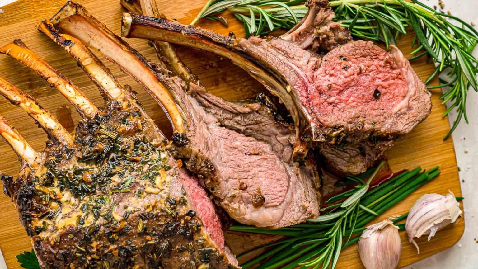 Sliced herb-crusted rack of lamb on a wooden board, garnished with fresh rosemary and garlic cloves.