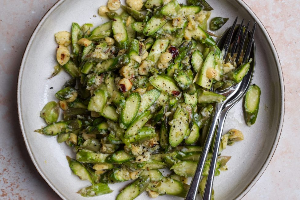 A plate of sliced asparagus mixed with chopped nuts and a light seasoning, served with a fork and spoon on a white dish.