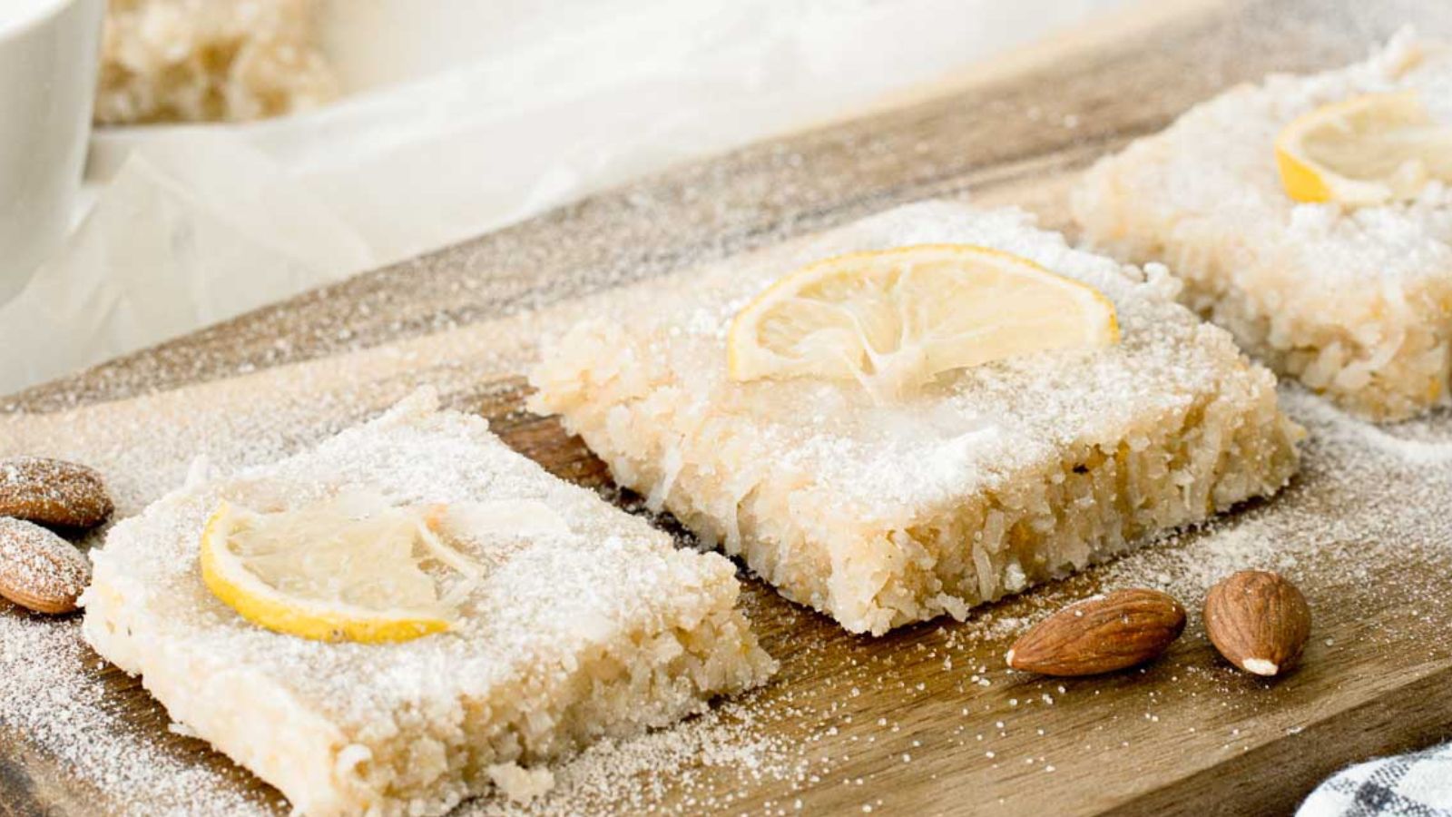 Three almond pastries topped with lemon slices and powdered sugar are arranged on a wooden board, with whole almonds scattered nearby.