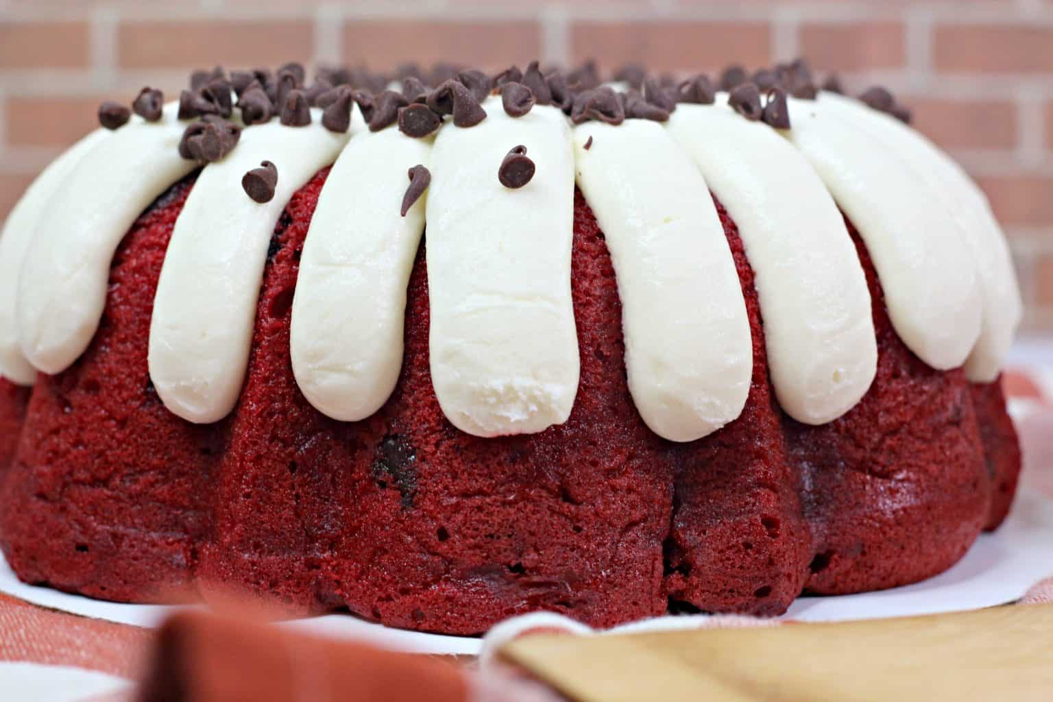 Red velvet bundt cake topped with thick white icing drizzles and sprinkled with chocolate chips, placed on a table with a brick wall background.