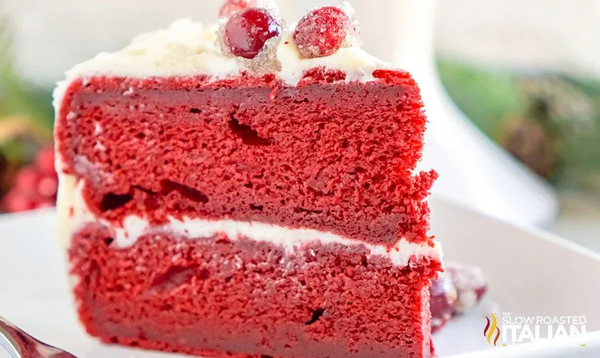 A slice of red velvet cake with white frosting and sugared cranberries on top, served on a white plate.