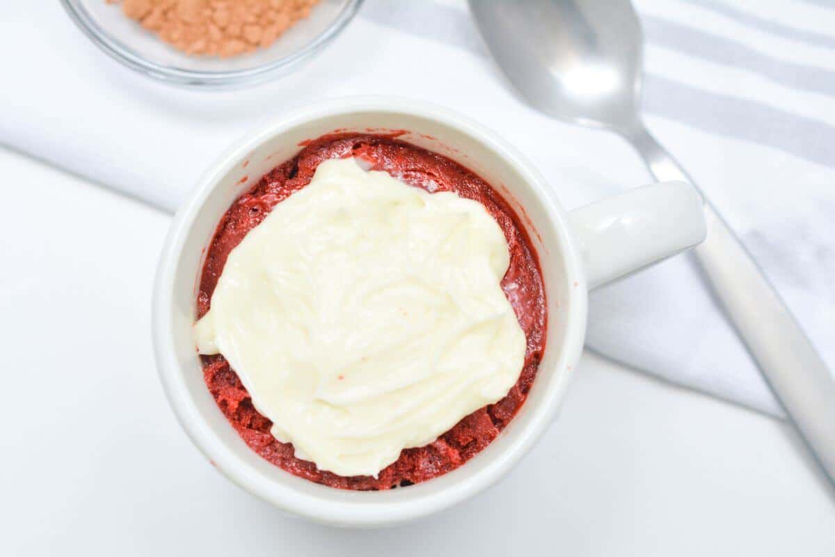 A white mug filled with red velvet cake topped with cream cheese frosting, placed on a white surface near a spoon and a bowl of cocoa powder.