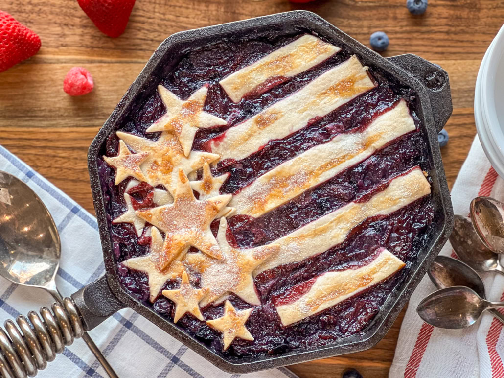 A berry pie in a skillet is decorated with pastry stars and stripes to resemble the American flag, sitting on a wooden table with berries and utensils nearby.